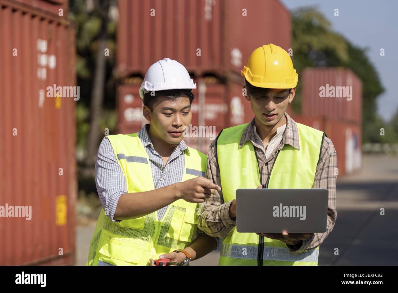 Zwei junge Ingenieure im Bereich Sicherheitsausrüstung analysieren Daten auf einem Laptop, während sie im Freien Logistikstrategien diskutieren Stockfoto