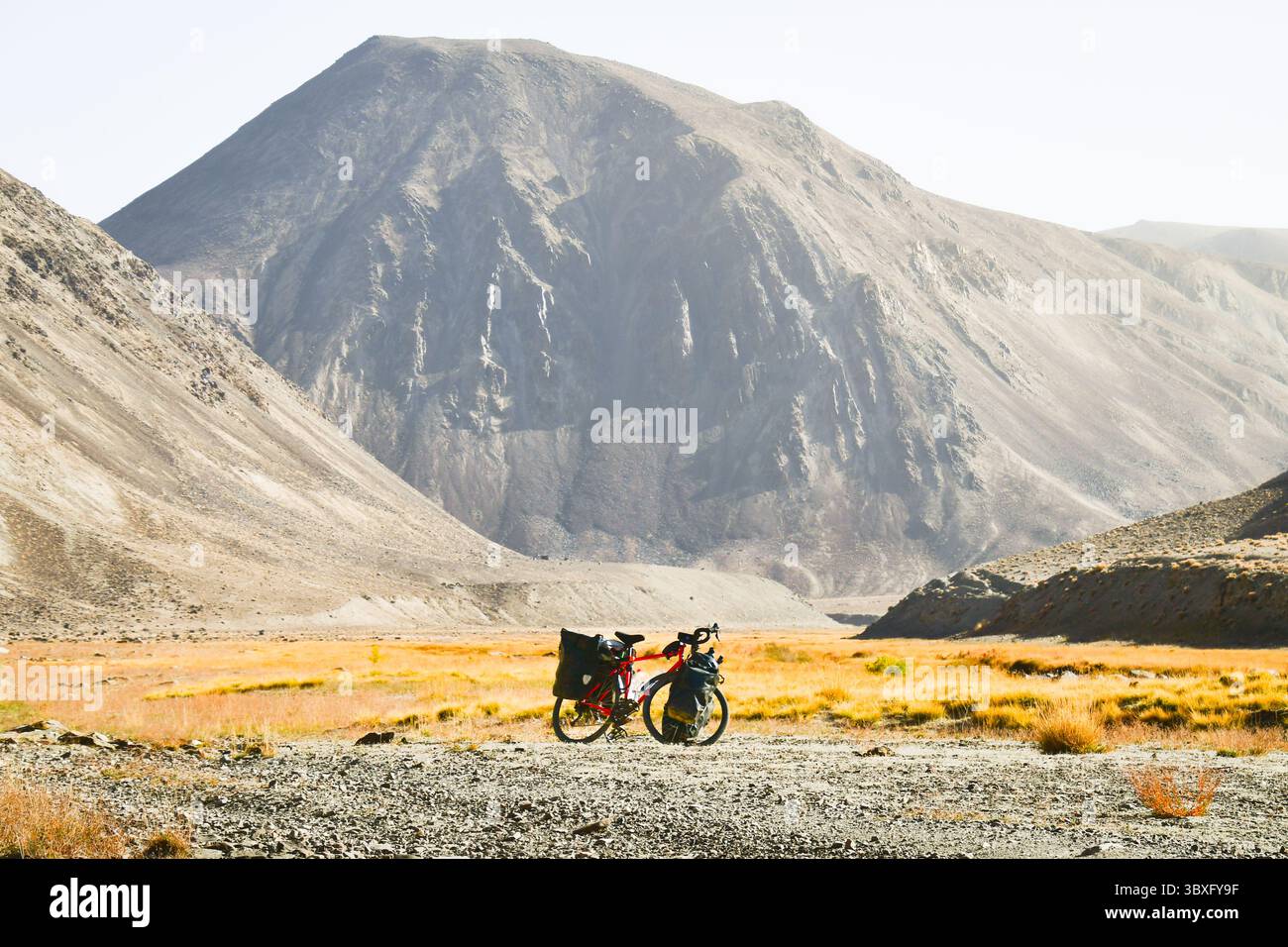 Rotes Tourenrad mit schwerem Gepäck in vier Gepäcktaschen steht an der Schotterstraße, umgeben von wilder, ariden Pamir-Bergen, alpiner Natur Stockfoto
