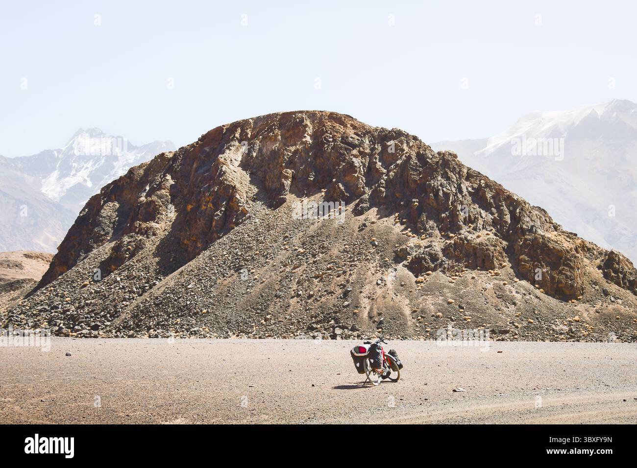 Rotes Tourenrad mit schwerem Gepäck in vier Gepäcktaschen steht an der Schotterstraße, umgeben von wilder, ariden Pamir-Bergen, alpiner Natur Stockfoto
