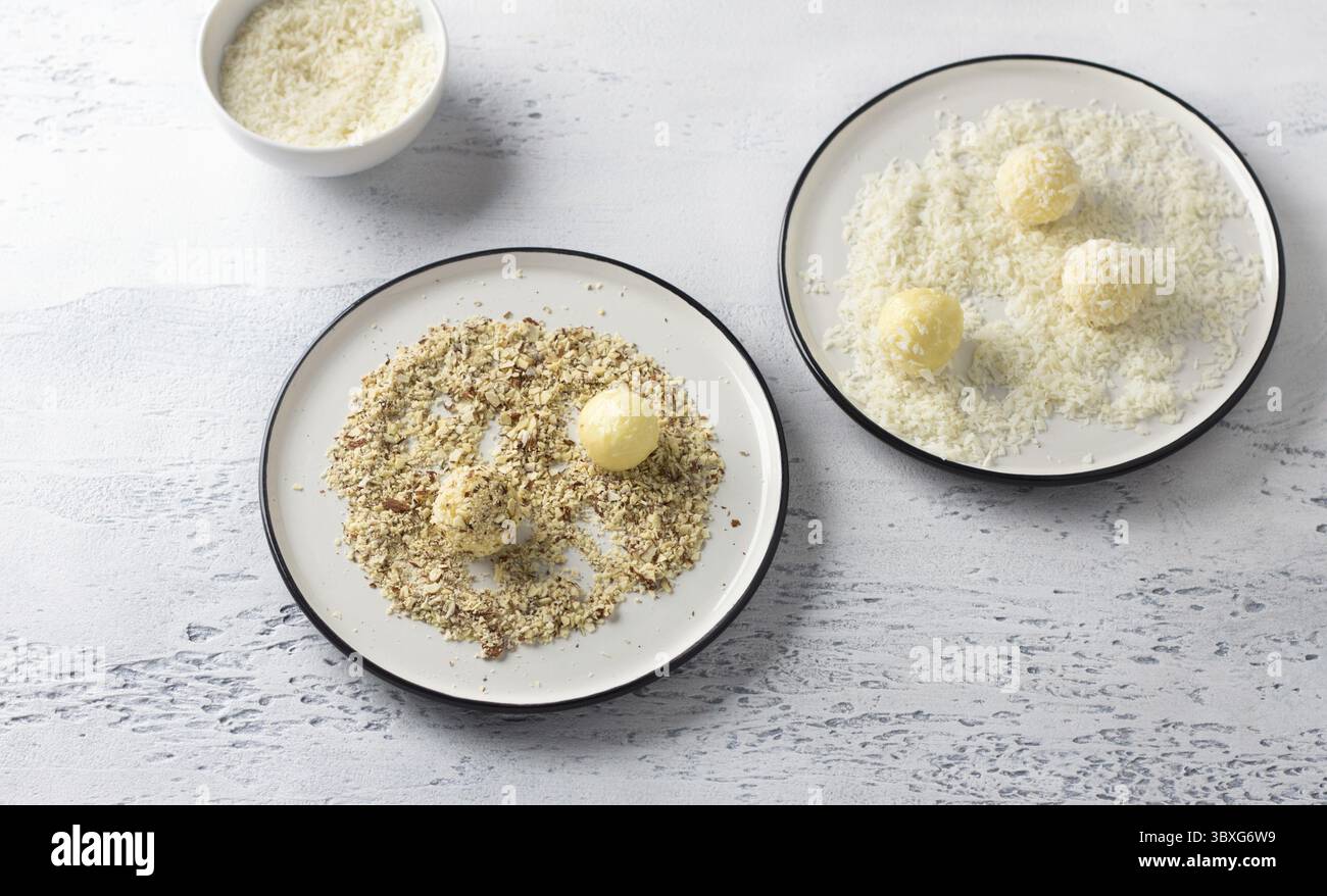 Essen, weiße Schokoladentrüffel in Kokosflocken gerollt und zerstoßene Nüsse auf einem weißen Teller, Blick von oben. Kochstufe mit köstlichen Pralinen Stockfoto