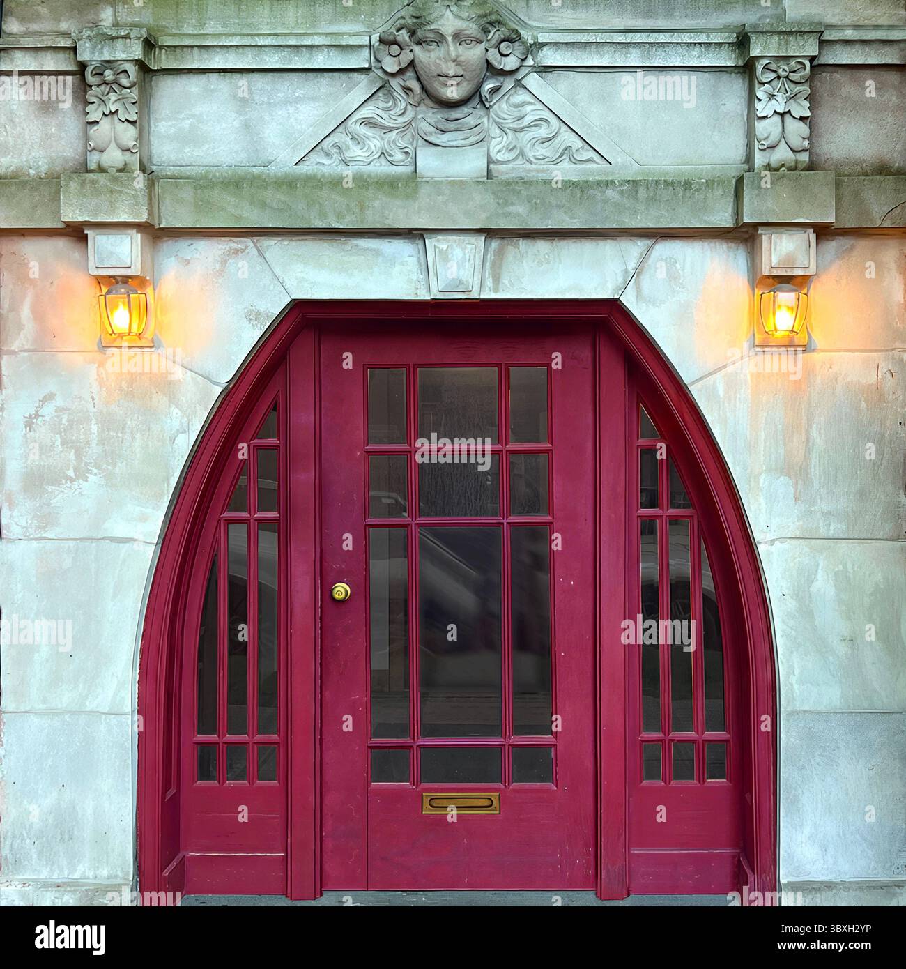 Rote Bogentür, eingerahmt von geschnitztem Stein und leuchtenden Laternen, gekrönt von einem plastischen Gesichtsdetail – ein kunstvoller Chicagoer Eingang mit gotischem Charme. - Smartphone-aufgenommenes Stockfoto