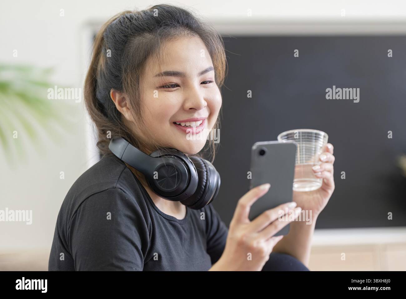 Eine junge Frau genießt zu Hause eine Yoga-Sitzung, trägt Kopfhörer und benutzt ein Smartphone, hält ein Glas Wasser und lächelt Stockfoto