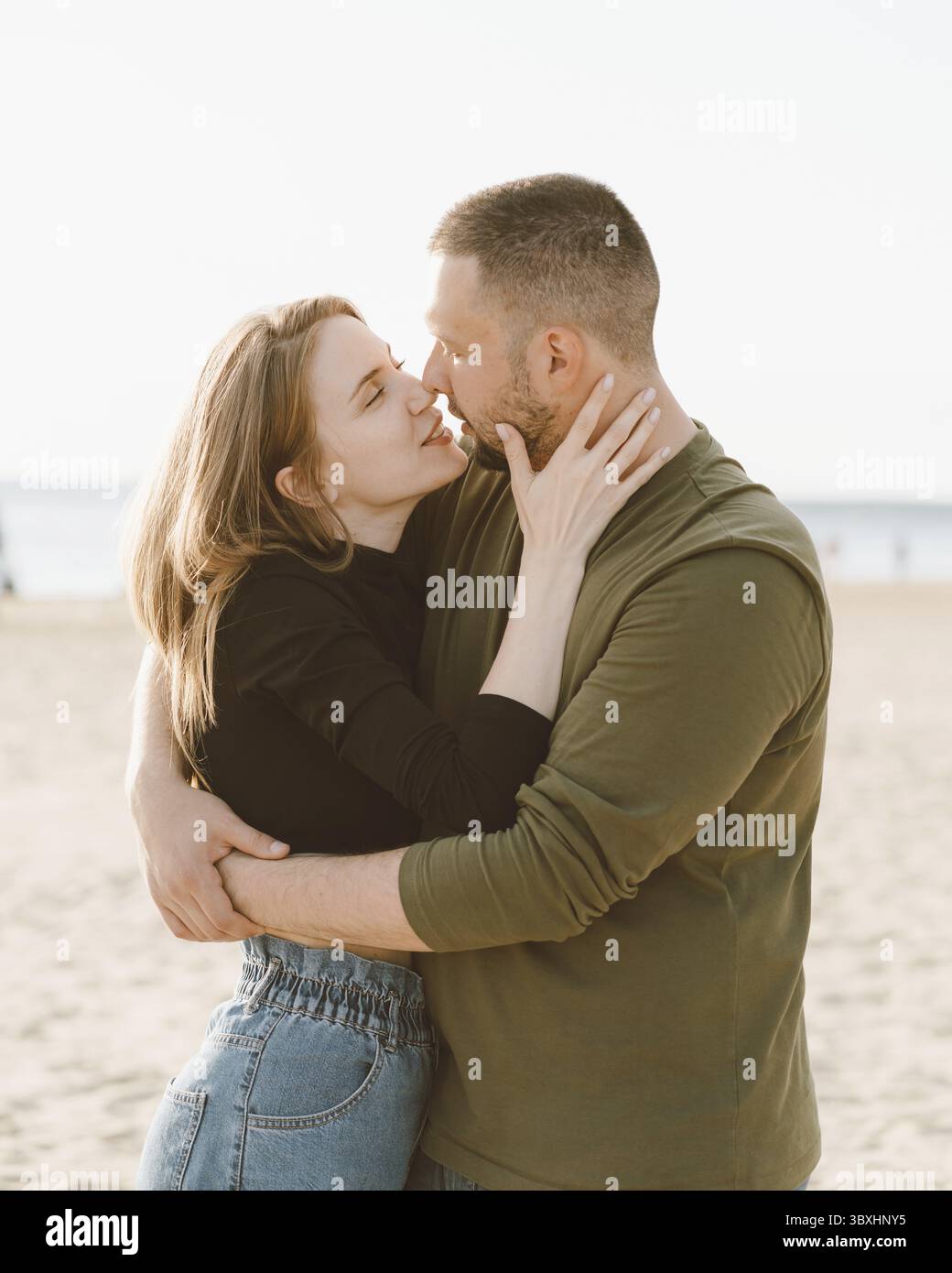 Junges erwachsenes Paar, das am Strand steht und sich ansieht. Hübscher, lächelnder Mann, der eine schöne Frau voller Liebe umschließt. Millennial männlich und weiblich p Stockfoto