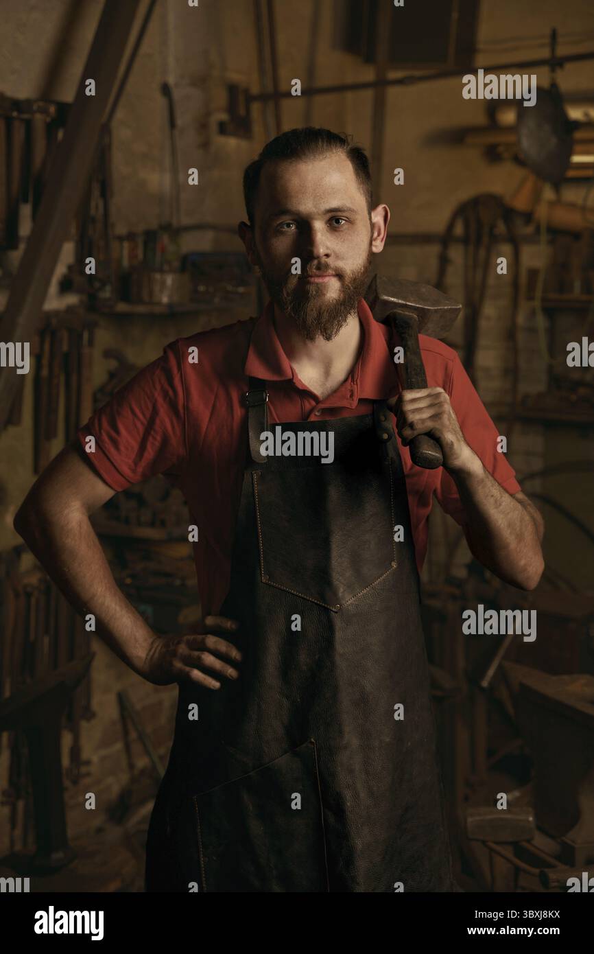 Professioneller Schmied im Werkstattporträt. Ernster junger europäischer bärtiger Mann mit Hand auf Hüften hält Hammer. Idee der handgefertigten Metallarbeit. Konzept von h Stockfoto