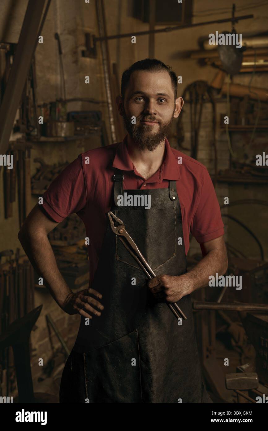 Professioneller Schmied im Schmiedeporträt. Positiver junger europäischer bärtiger Mann mit Hand auf Hüften hält eine Eisenzange. Idee der handgefertigten Metallarbeit. Konzept o Stockfoto