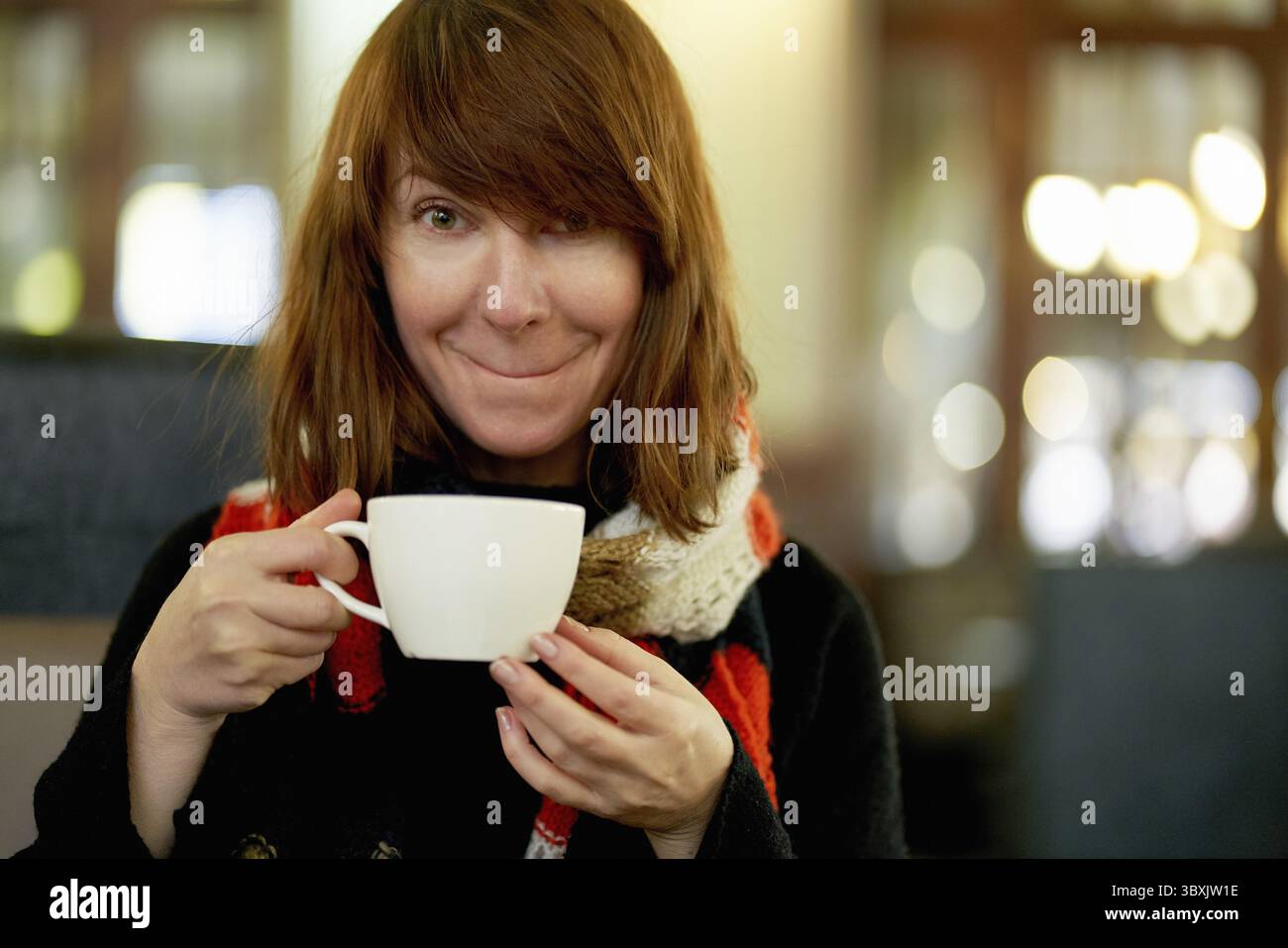 Das schöne Weibchen trinkt Kaffee im Café. Reife Frau mit langen Haaren schaut in die Kamera, trinkt Kaffee und lächelt hübsch. Kopierbereich. Großes weiß Stockfoto