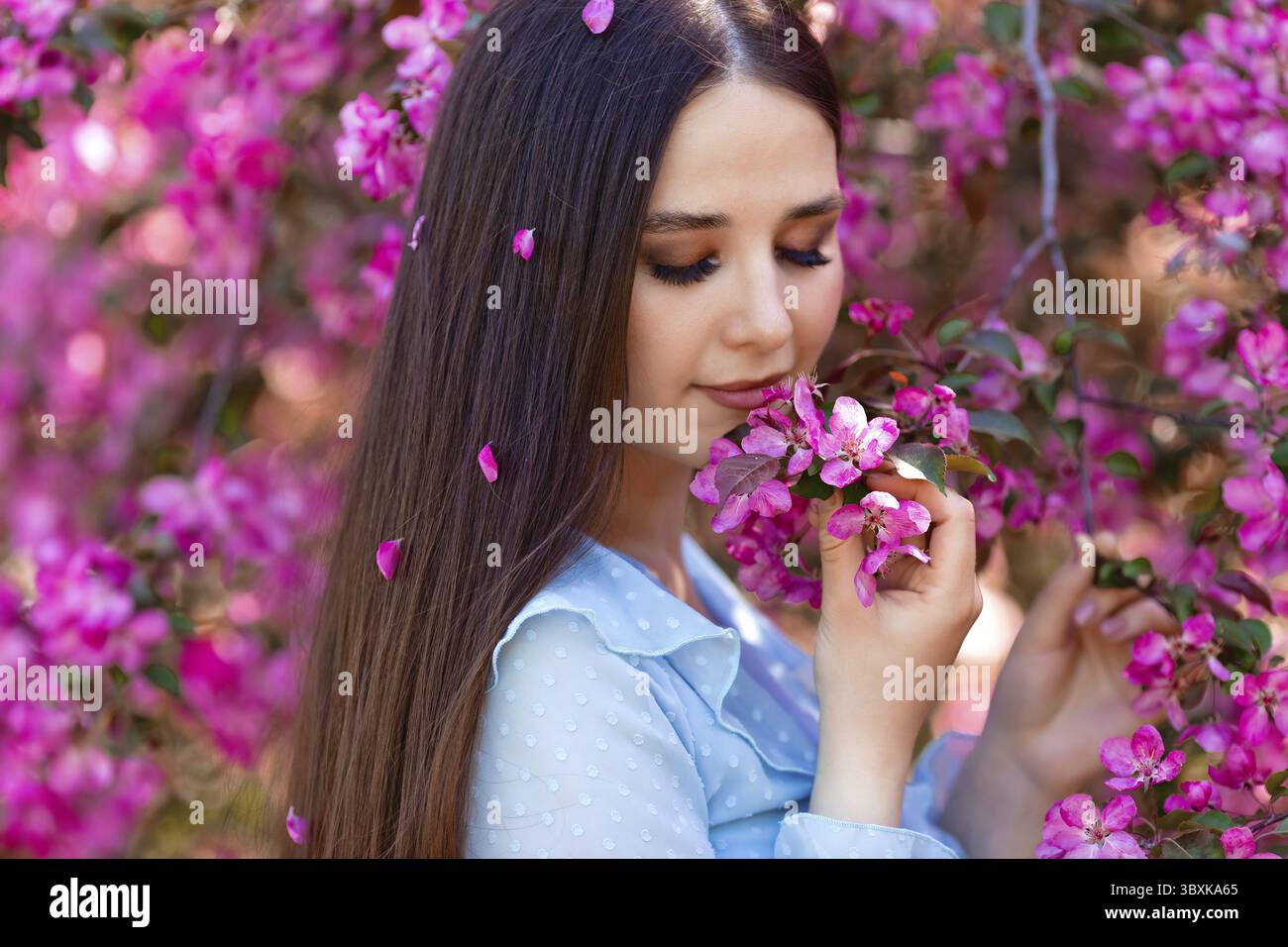Ein schönes, brünettes Mädchen mit rosa Blüten in ihren langen Haaren steht an einem sonnigen Tag neben einem rosa blühenden Apfelbaum Stockfoto