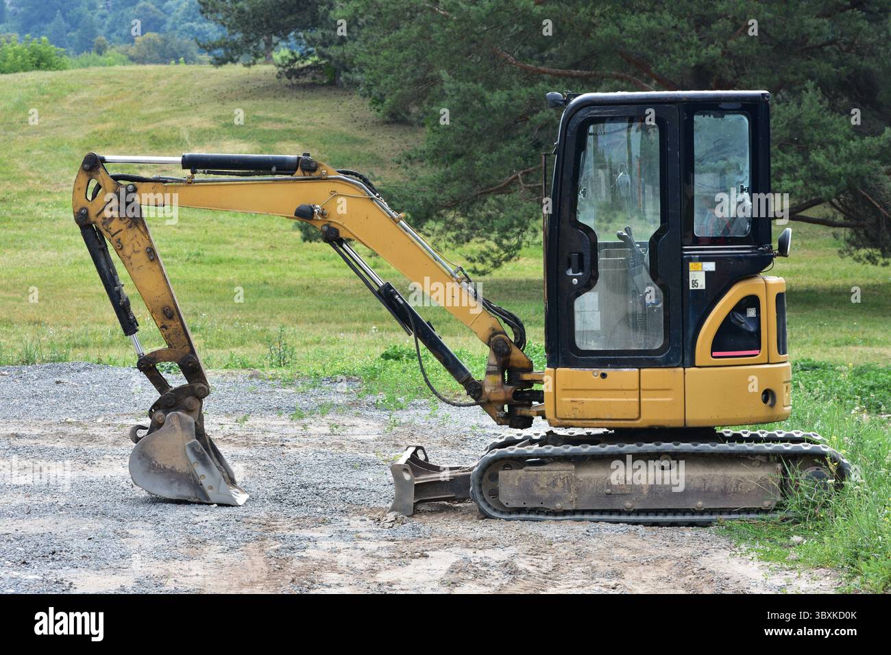 Kompakter gelber caterpillar-Bagger steht auf Schotter in der Nähe von Grasfeldern. Baumaschinen für Graben, Erdarbeiten und kleine Projekte. Stockfoto
