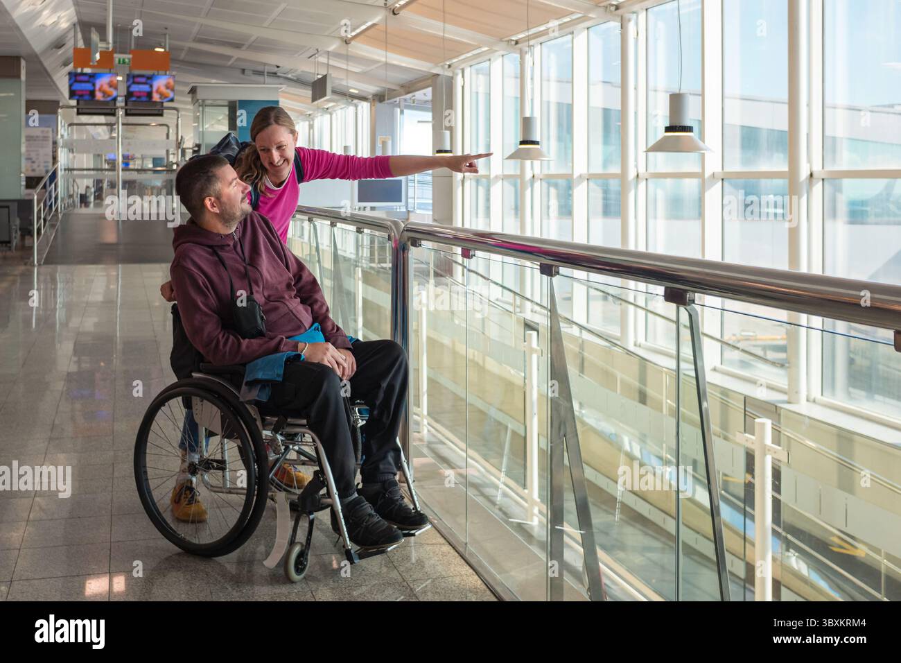 Mann mit Behinderung, der Rollstuhl mit seiner weiblichen persönlichen Assistentin am Flughafen benutzt. Sie schauen aus den großen Fenstern, während sie sich auf ihre vorbereiten Stockfoto