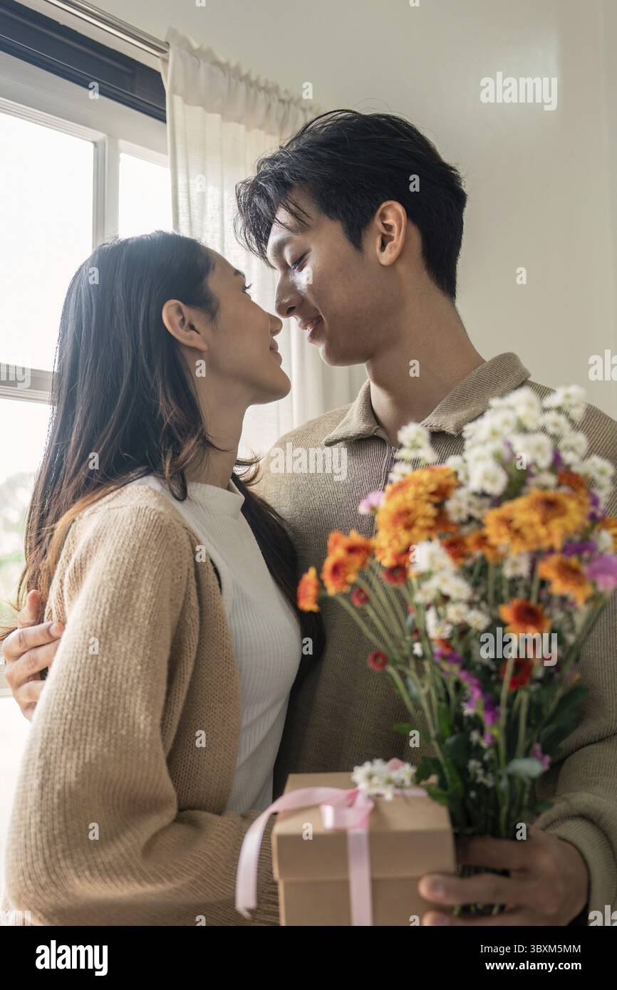 Romantisches junges asiatisches Paar, das sich mit Blumen umschließt und im Wohnzimmer zu Hause lächelt. Verlieben Sie sich. Valentinstag-Konzept Stockfoto