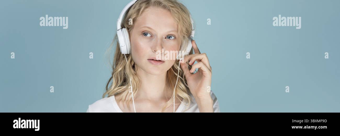 Langes Banner mit Porträt einer jungen Frau, die Musik über Kopfhörer hört, auf farbneutraler Aqua Menthe Wand. Ziemlich ernste, clevere blonde Frau Stockfoto