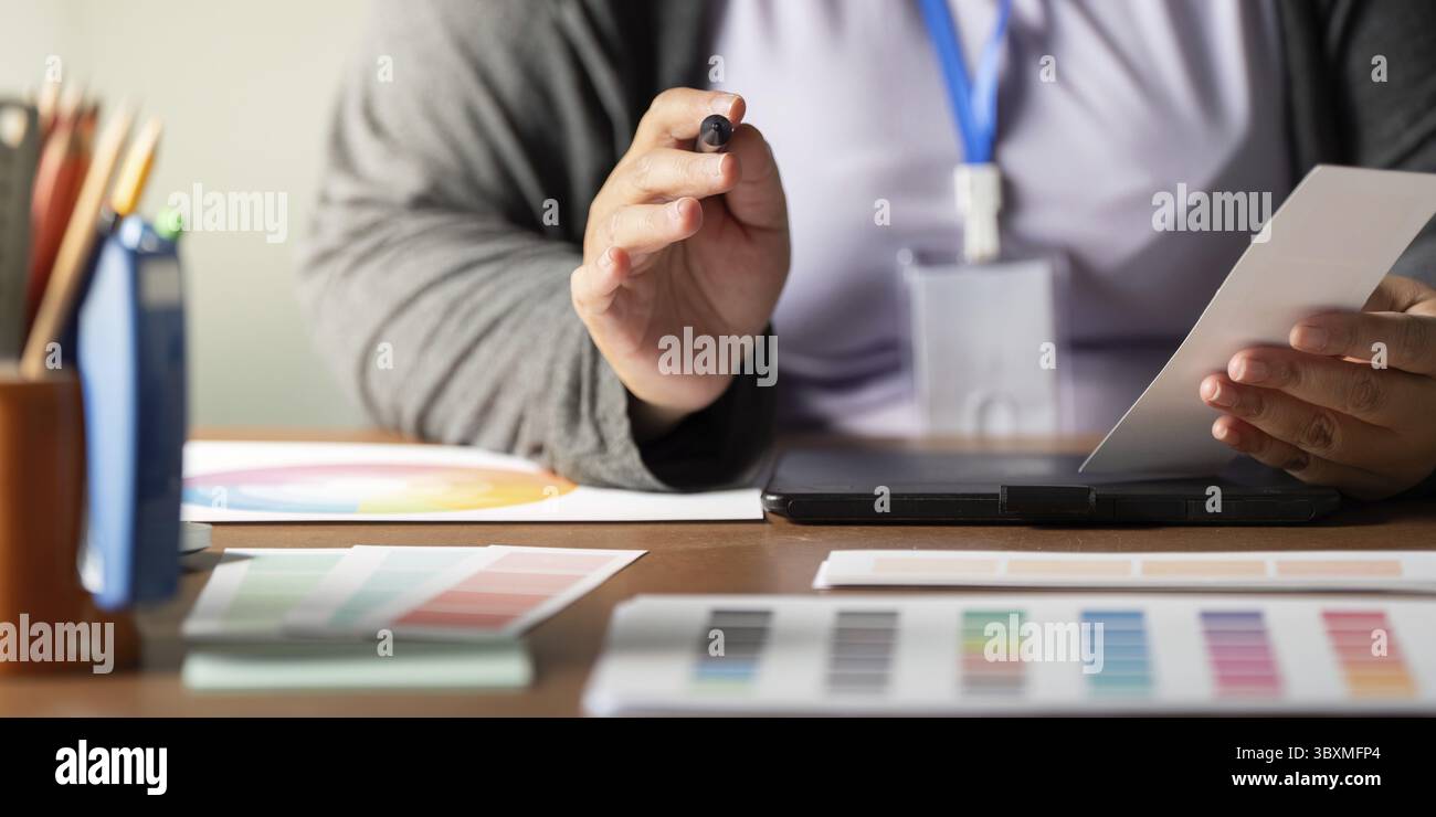 Ein Grafikdesigner diskutiert Designkonzepte und hält dabei einen Marker, umgeben von Farbpaletten und Designmaterialien, auf einem eleganten Schreibtisch Stockfoto