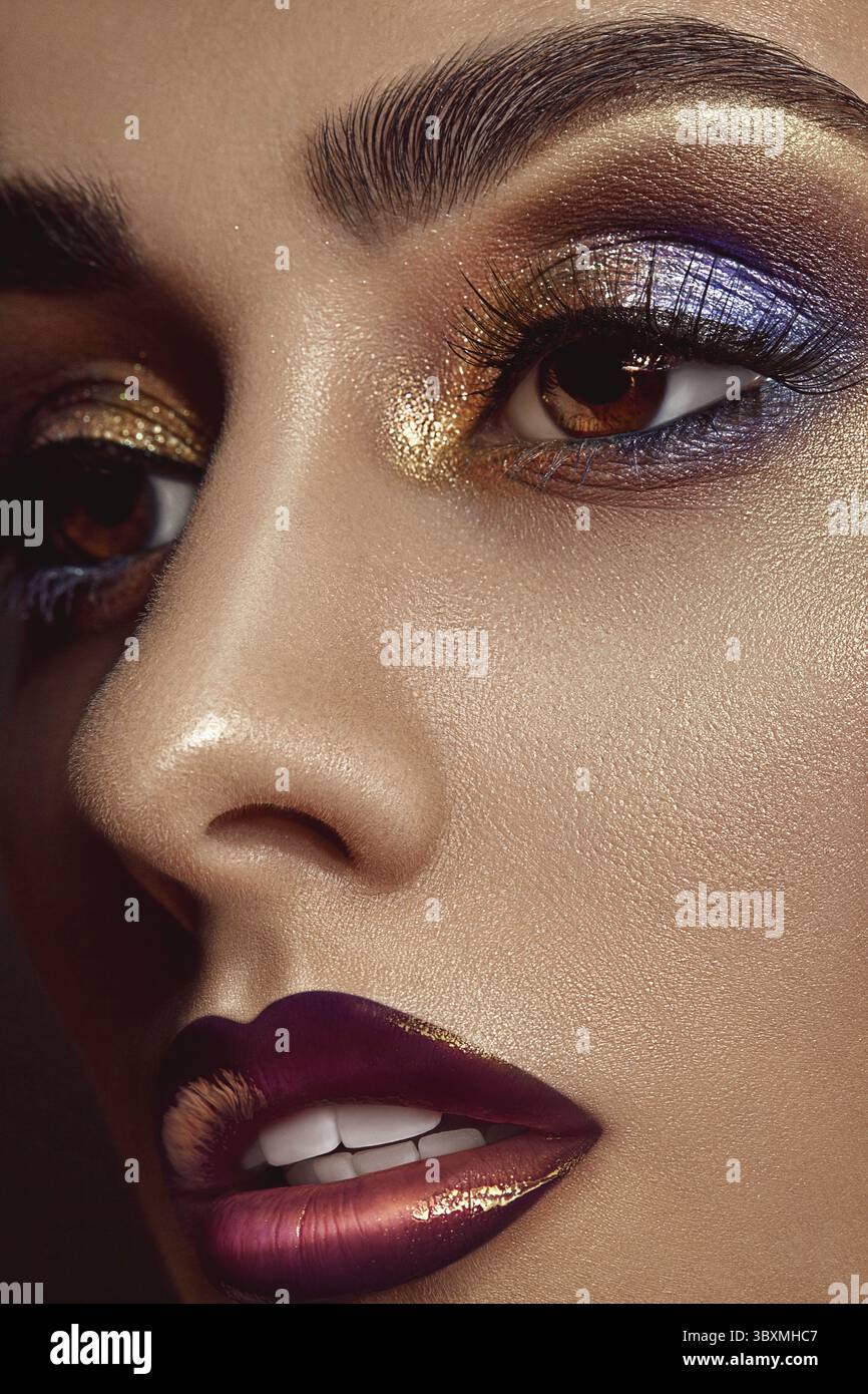 Brunette-Dame mit luxuriösem Make-up und perfekter Haut schaut weg. Blaue und goldene Augenschatten, lange falsche Wimpern, glänzende weinrote Lippen und weißes T-Shirt Stockfoto