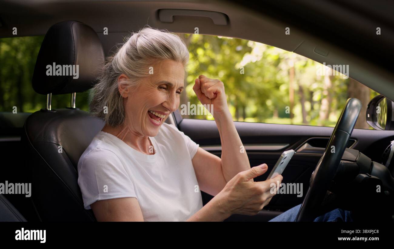 Fröhliche kaukasische reife Frau ältere ältere ältere Frau Rentner Auto Fahrer im Auto mit Handy spielen Spiel Ballen Faust Stockfoto