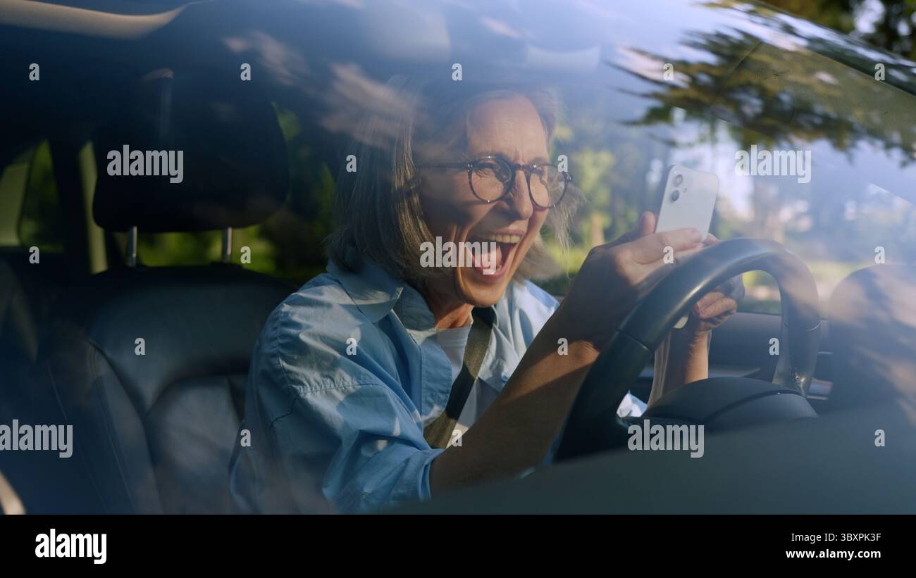 Aufgeregt schockiert überrascht kaukasische ältere ältere Frau weibliche ältere Fahrer Auto Besitzer im Auto mit Handy-App gewinnen lesen großartig Stockfoto