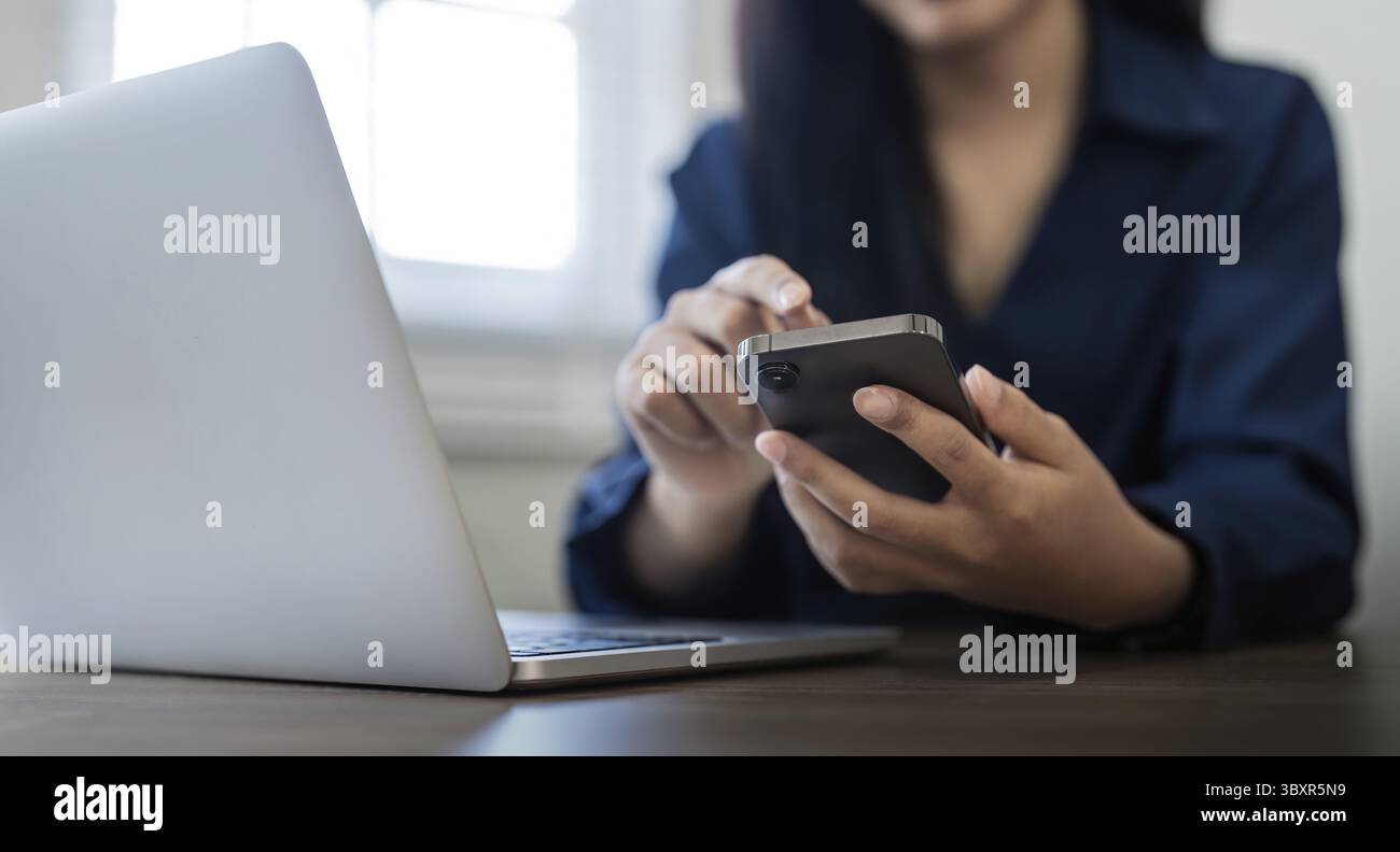 Eine junge Geschäftsfrau am Telefon im Büro. Geschäftsfrau, die am Telefon SMS schreibt und am Laptop arbeitet. Hübsche junge Geschäftsfrau, die auf der Arbeitsplatte sitzt Stockfoto
