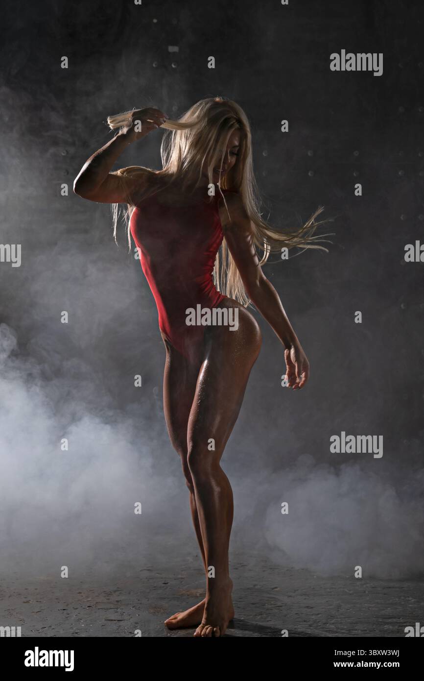 Die geschickte blonde Athletin steht selbstbewusst und betont ihren Körper und ihre anmutige Bewegung in rotem Outfit, umgeben von Nebel in dramatischer Umgebung Stockfoto