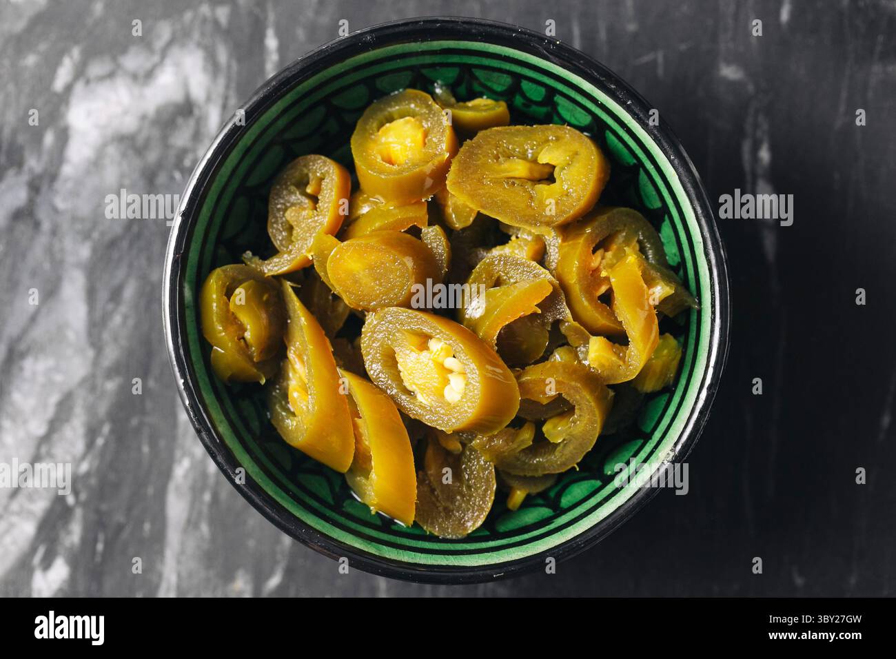 Eingelegte Jalapeno-Chilischeiben in Keramikschale. Scharfe grüne Paprika auf dunklem Marmorhintergrund. Würzig konservierte Gemüsezusatz zum Kochen oder Snack Stockfoto