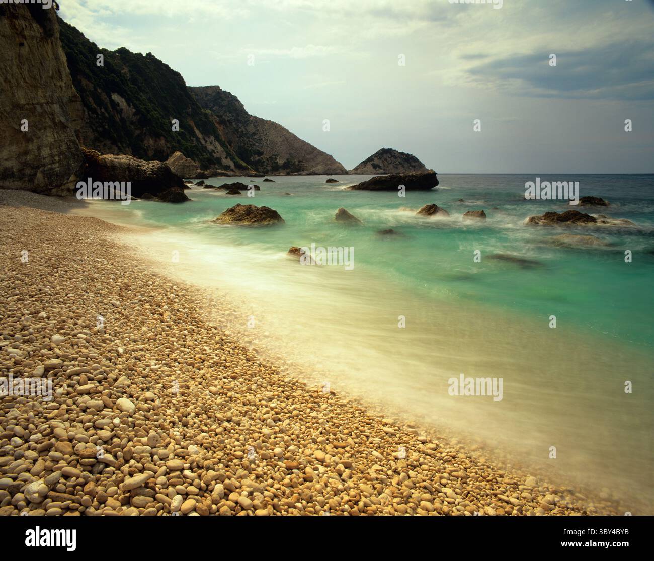 Das Mittelmeer stürzt auf die Kieselsteine und Felsen des Petani-Strandes, während die Sonne durch die Sommersturmwolken Anfang Juni kämpft Stockfoto