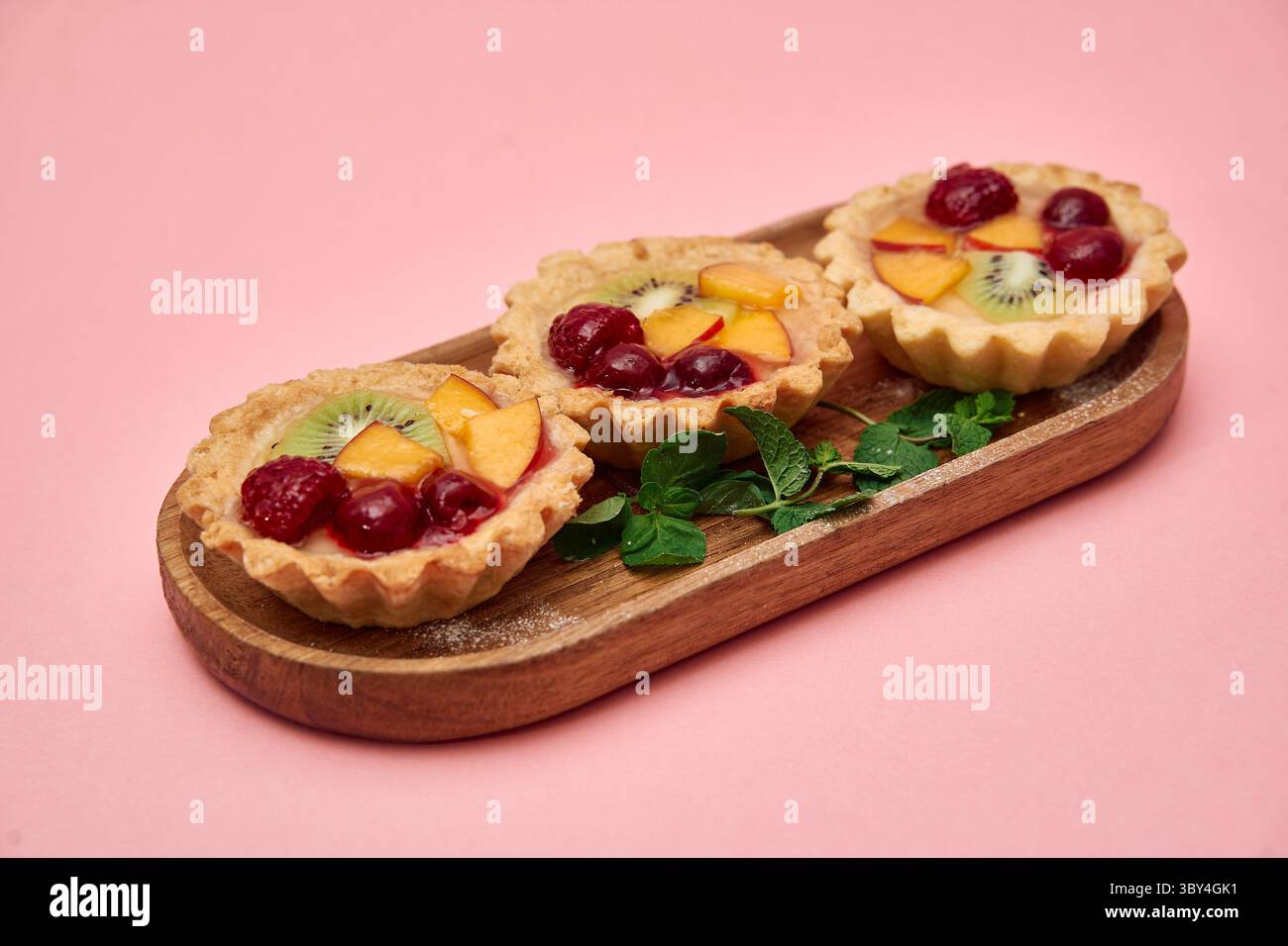 Drei köstliche Obsttörtchen, garniert mit Kirschen, Kiwis und Pfirsichen, sind auf einer Holzplatte angeordnet, ergänzt durch frische Minzblätter auf rosa Stockfoto