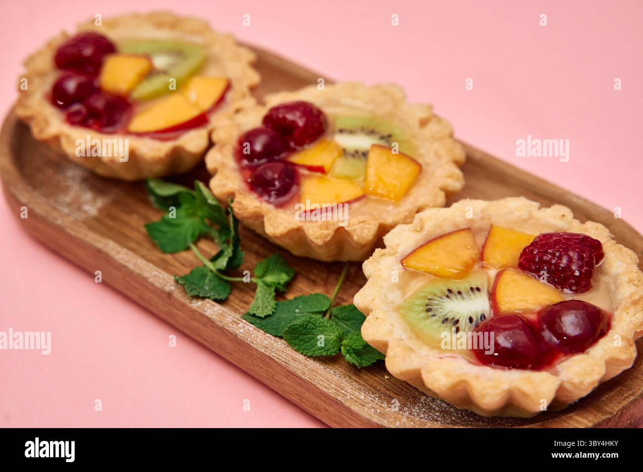 Leckere Mini-Obstkuchen mit Kiwi, Pfirsich, Himbeere und Kirsche, garniert mit Minze auf einem Holztablett, bieten eine lebendige und appetitliche Atmosphäre Stockfoto