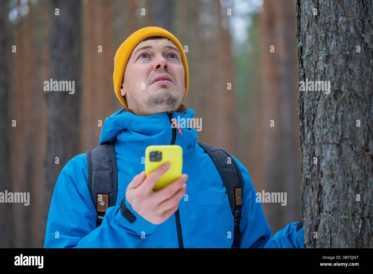 Männlicher Wanderer in blauer Jacke und gelber Beanie, der in einem Winterwald nach einem Mobilfunksignal sucht und ein Smartphone in der Hand hält Stockfoto