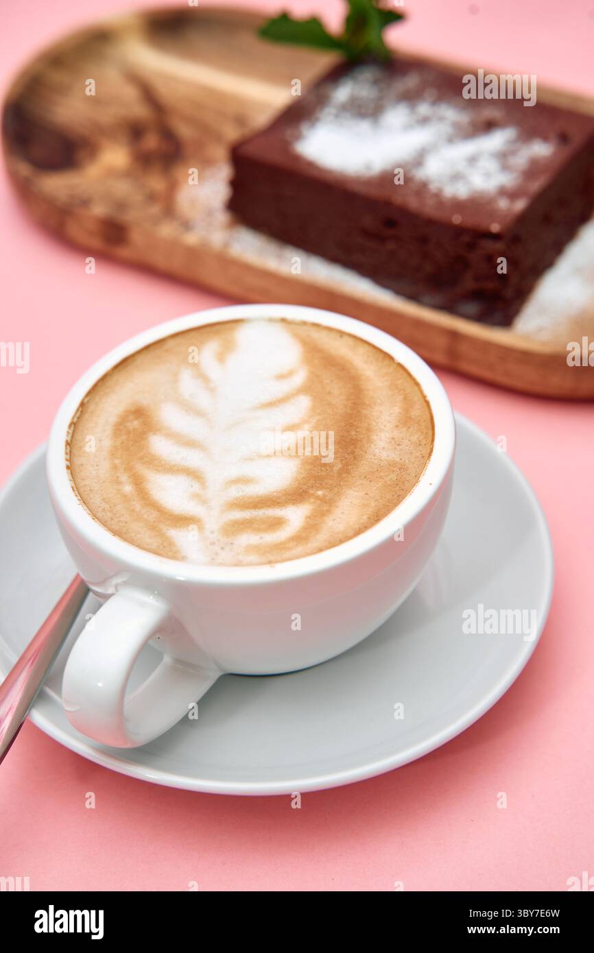 Ein cremiger Latte mit zarter Blattkunst zusammen mit einem dekadenten Schokoladenkuchen, wunderschön präsentiert auf einer rosafarbenen Oberfläche, schafft ein optisch ansprechendes und ein Stockfoto