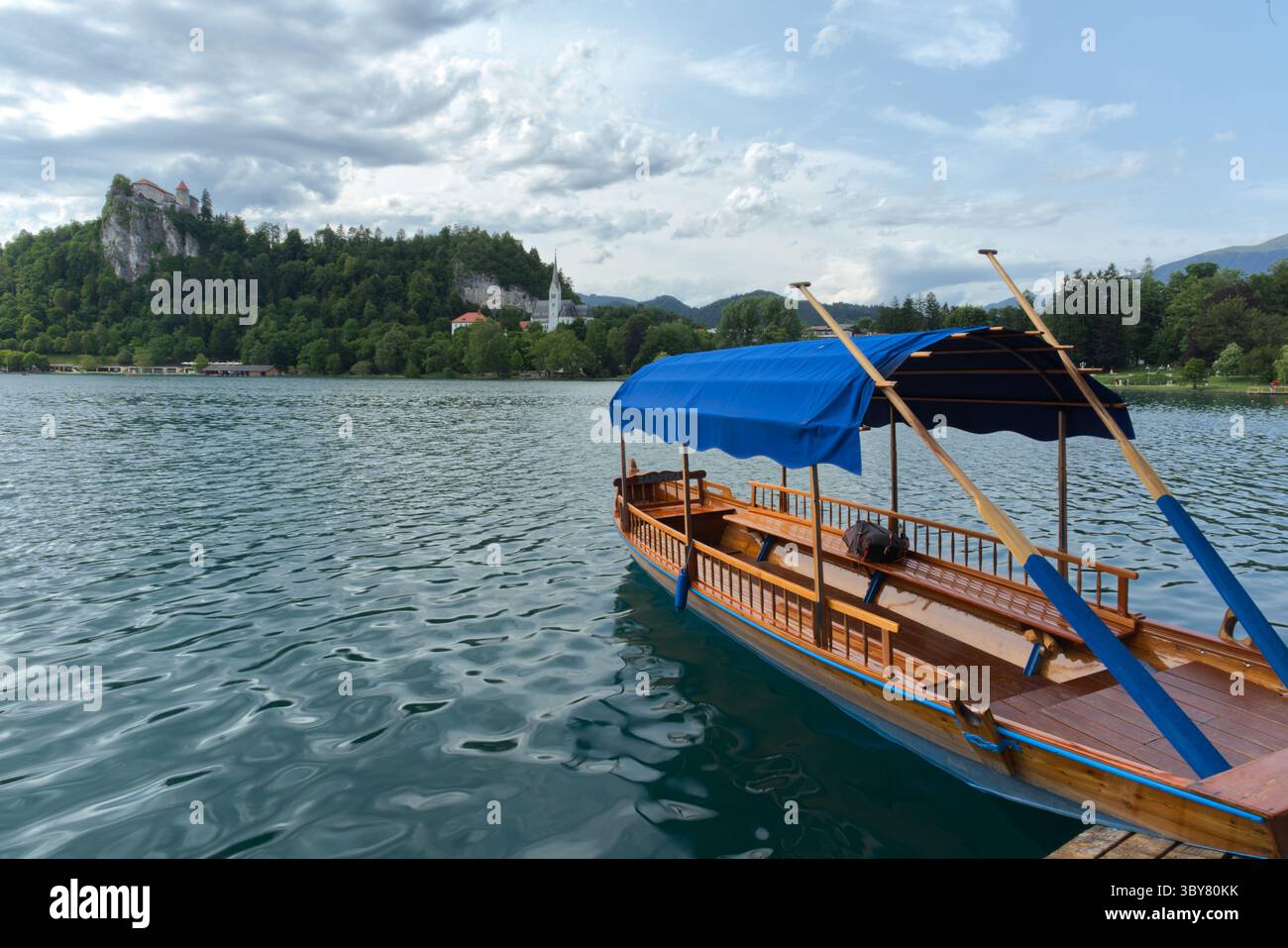 Ein traditionelles hölzernes Pletna-Boot ruht auf dem ruhigen Wasser des Bleder Sees, mit dem berühmten Schloss von Bled auf einer Klippe und der Kirche Stockfoto