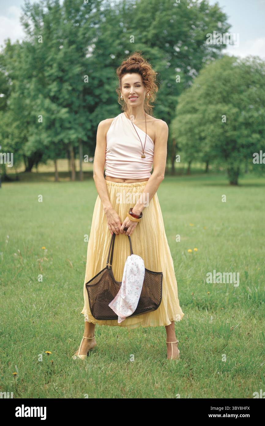 Eine Frau steht in einem üppig grünen Park, trägt ein hellrosa Oberteil und einen fließenden gelben Rock. Sie hält eine dunkle Tragetasche mit einem weißen Tuch darin, umgeben Stockfoto