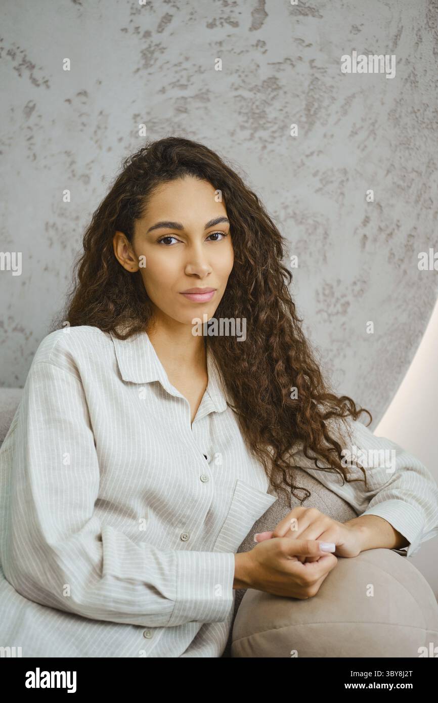 Eine junge Frau mit langen, lockigen Haaren liegt in einem modernen Raum auf einem Sofa. Sanftes Nachmittagslicht erfüllt den Raum und hebt ihre nachdenkliche Ex hervor Stockfoto
