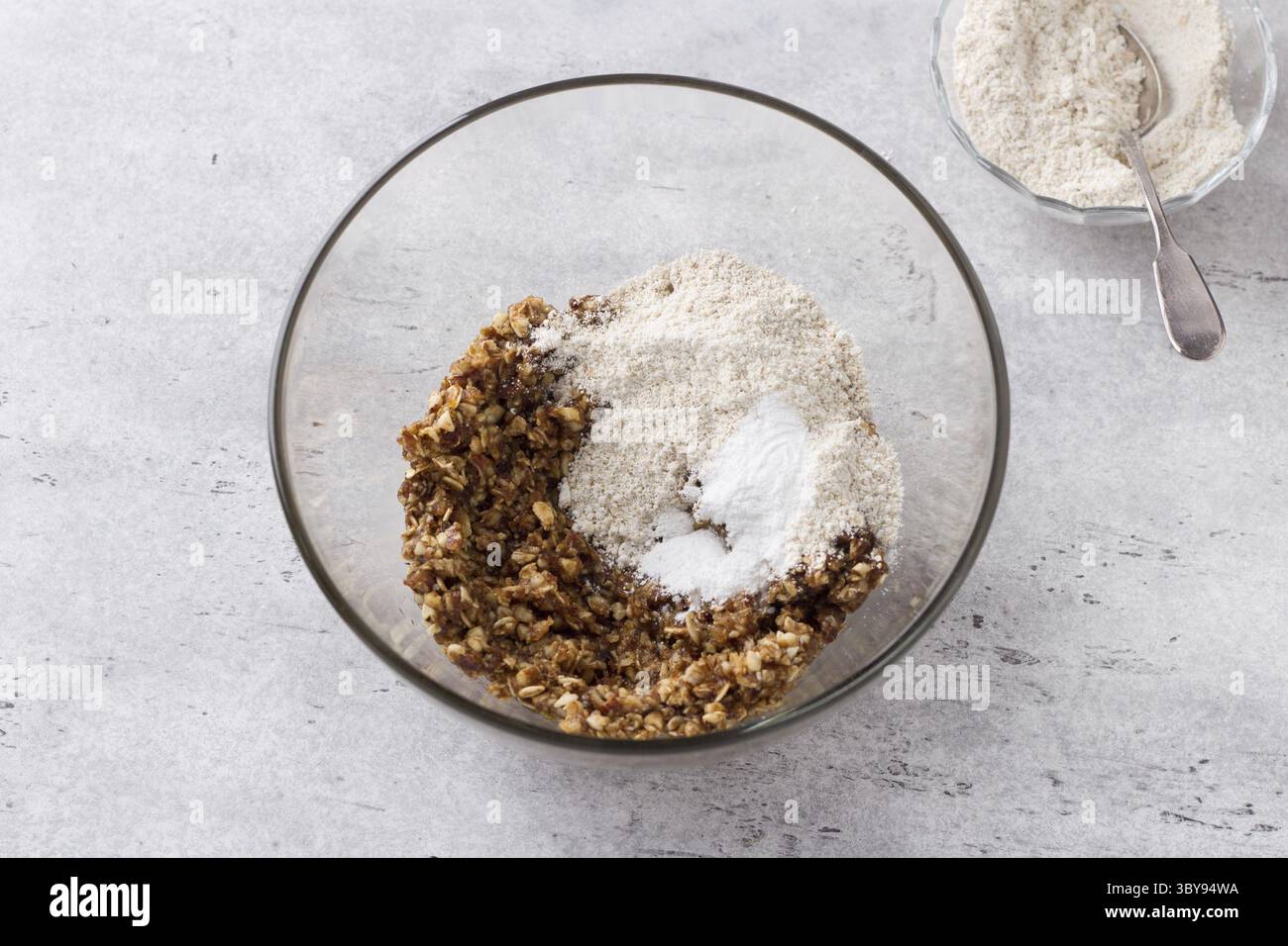 Essen, Kochen veganer gesunder Haferflockenkekse, Hinzufügen von Hafermehl und Backpulver auf einem grauen texturierten Hintergrund Stockfoto