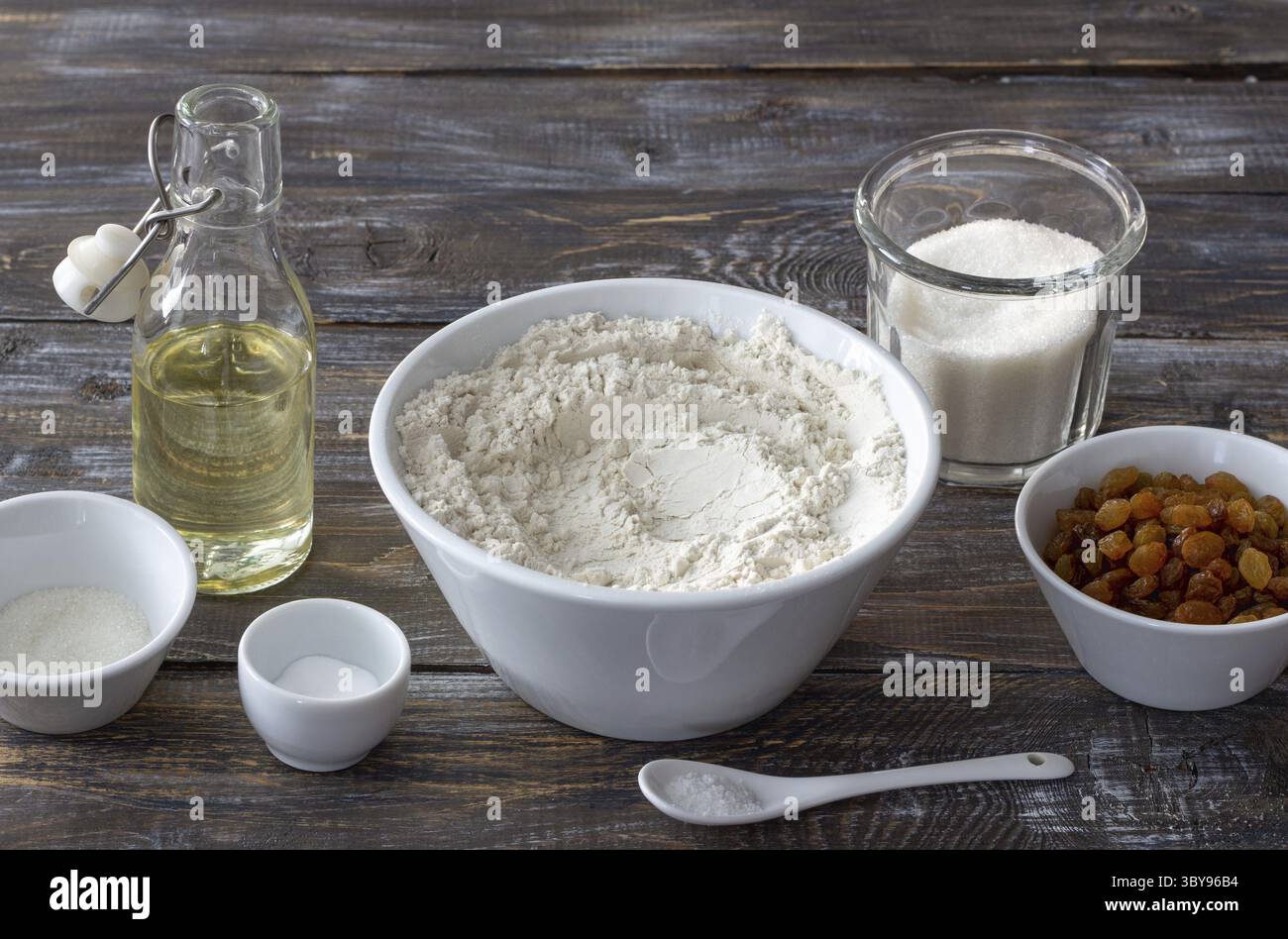 Lebensmittel, Backzutaten einschließlich Mehl, goldene Rosinen, Zucker, Öl, Soda, Vanillezucker und Salz in Schüsseln auf rustikalem Holztisch, für Rezeptkonzept Stockfoto