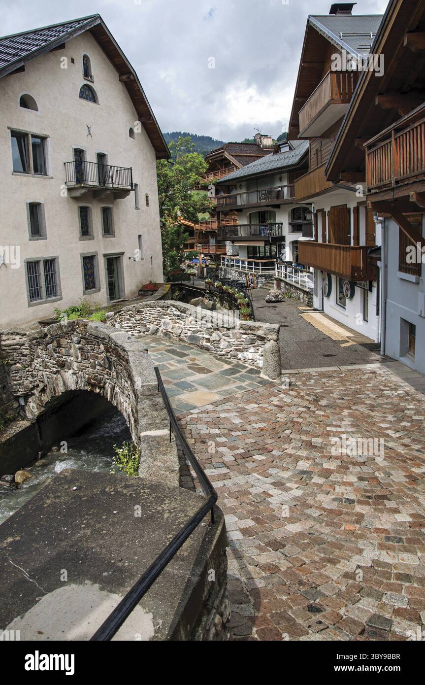Typische Gebäude in der Straße mit Kopfsteinpflaster, kleiner Brücke und Blick auf die Alpen an einem bewölkten Tag in Megeve. Ein berühmtes Skigebiet in der Haute-Savoie Stockfoto