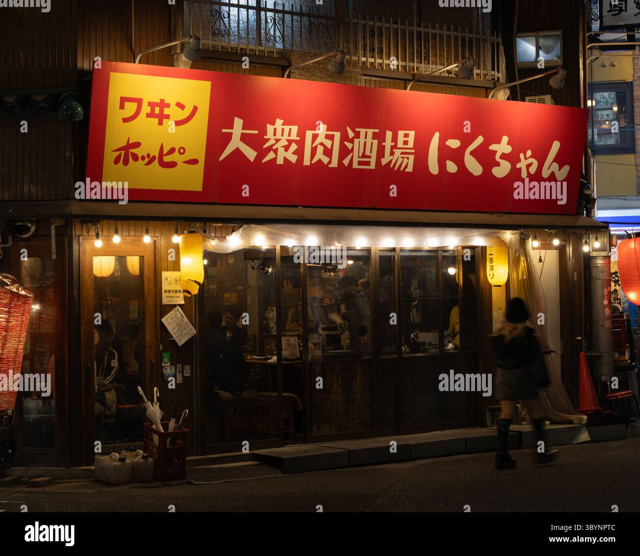 Das Bild zeigt ein japanisches Restaurant bei Nacht. Die Werbung ist rot und gelb und es gibt warme gelbe Lichter im Inneren des Restaurants. Th Stockfoto
