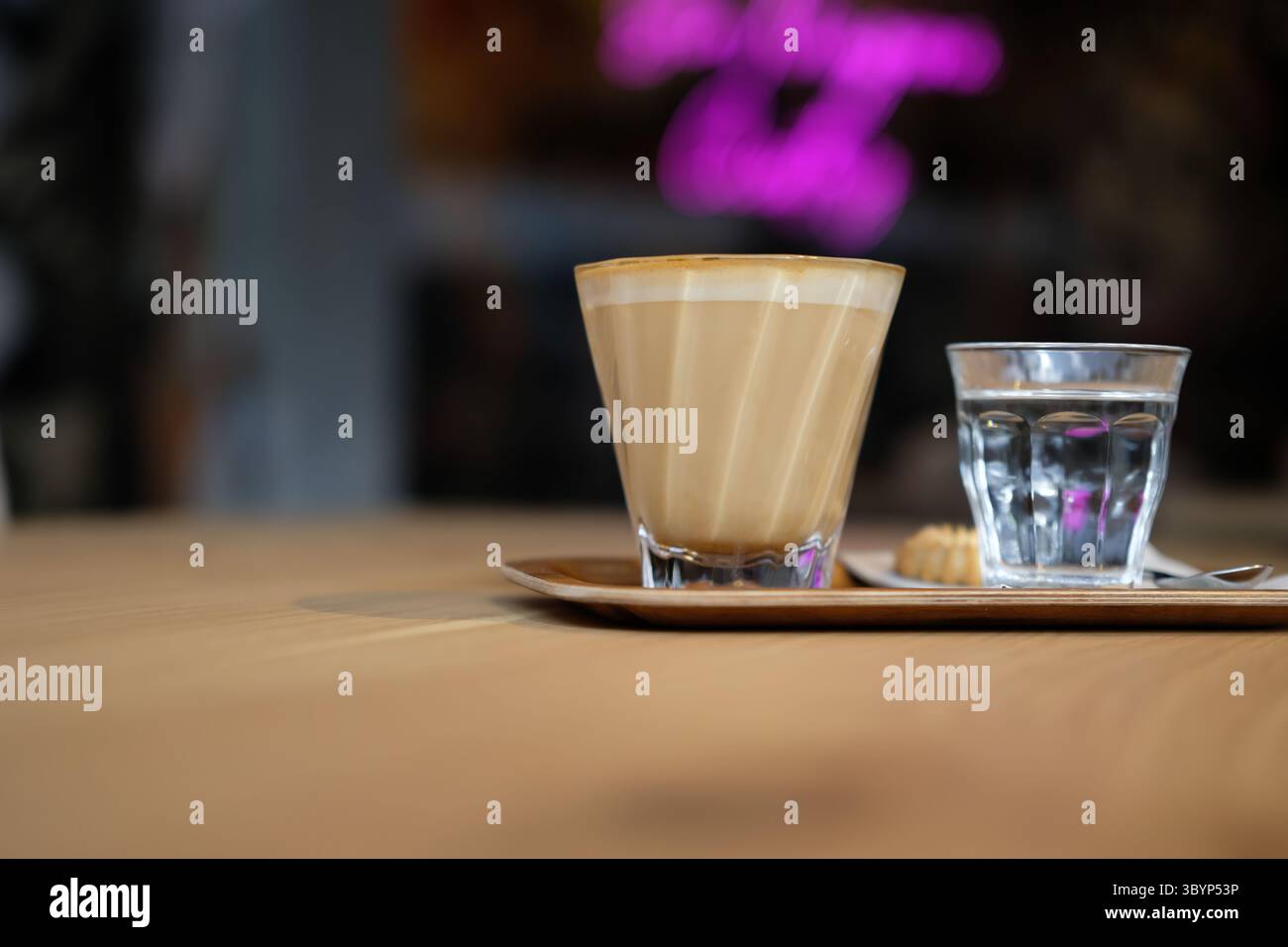 Flaches weiß mit Wasser und Keks auf Holztablett im modernen Café – gemütliche Kaffeepause, entspannender Lifestyle-Moment mit warmem Licht und weichem Bokeh. Stockfoto