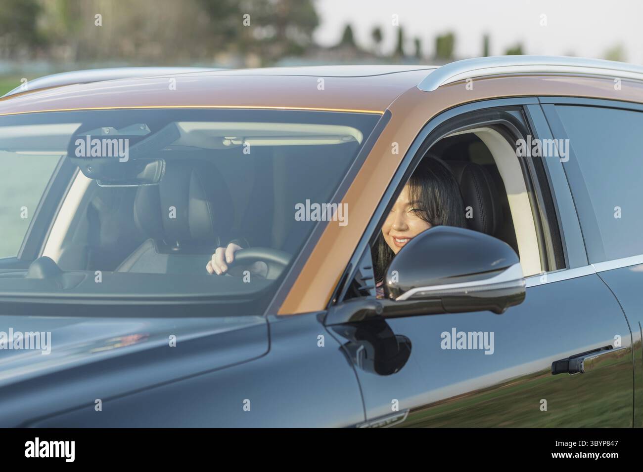 Eine Frau genießt ihre Zeit hinter dem Steuer eines modernen SUV und lächelt selbstbewusst. Das Fahrzeug steht in einer sonnigen Gegend in einer ruhigen Gegend. Stockfoto