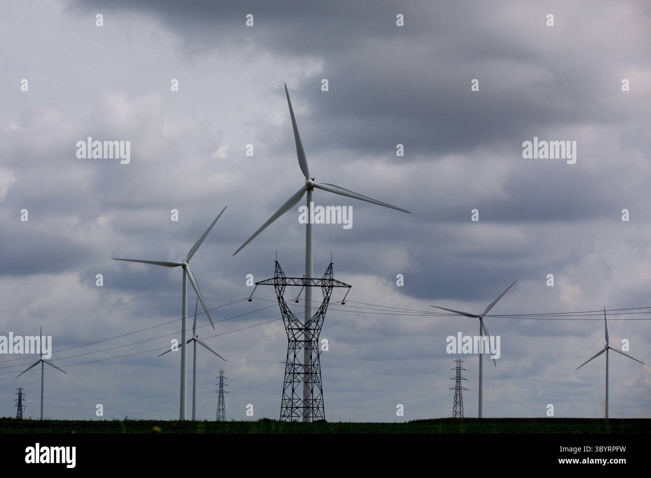 Luftpanorama von kaskadierenden Windturbinen und Strommasten unter dramatischen Wolken über Amsterdams flachem Polder, der die Konvergenz erneuerbarer Energien zeigt Stockfoto