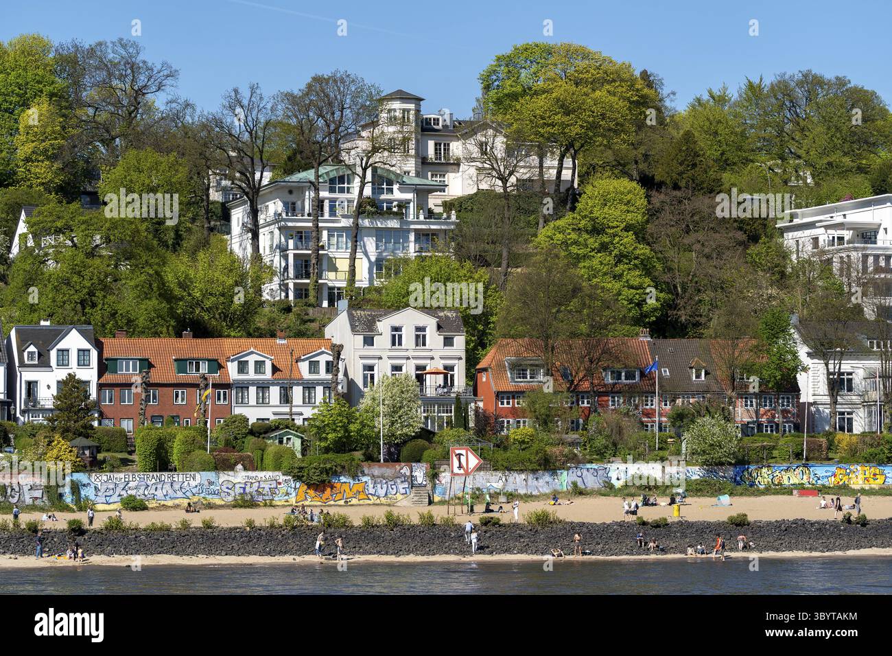 Blick auf den Elbstrand in Oevelgoenne mit zahlreichen Spaziergänger und Sonnenanbetern am Ufer, im Hintergrund historische Kapitänshäuser mit rotem Tiegel Stockfoto