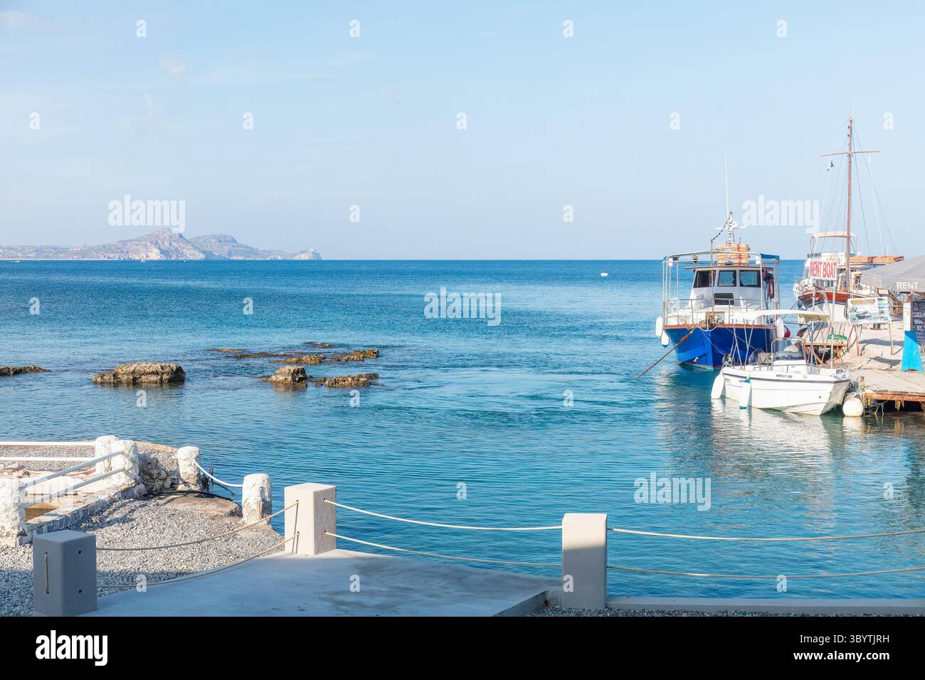 Der malerische Hafen mit lebhaften Booten, die auf dem ruhigen azurblauen Wasser ruhen, und felsige Formationen unterstreichen das ruhige Ambiente an der Küste. Griechenland, Insel Rhodos. Stockfoto