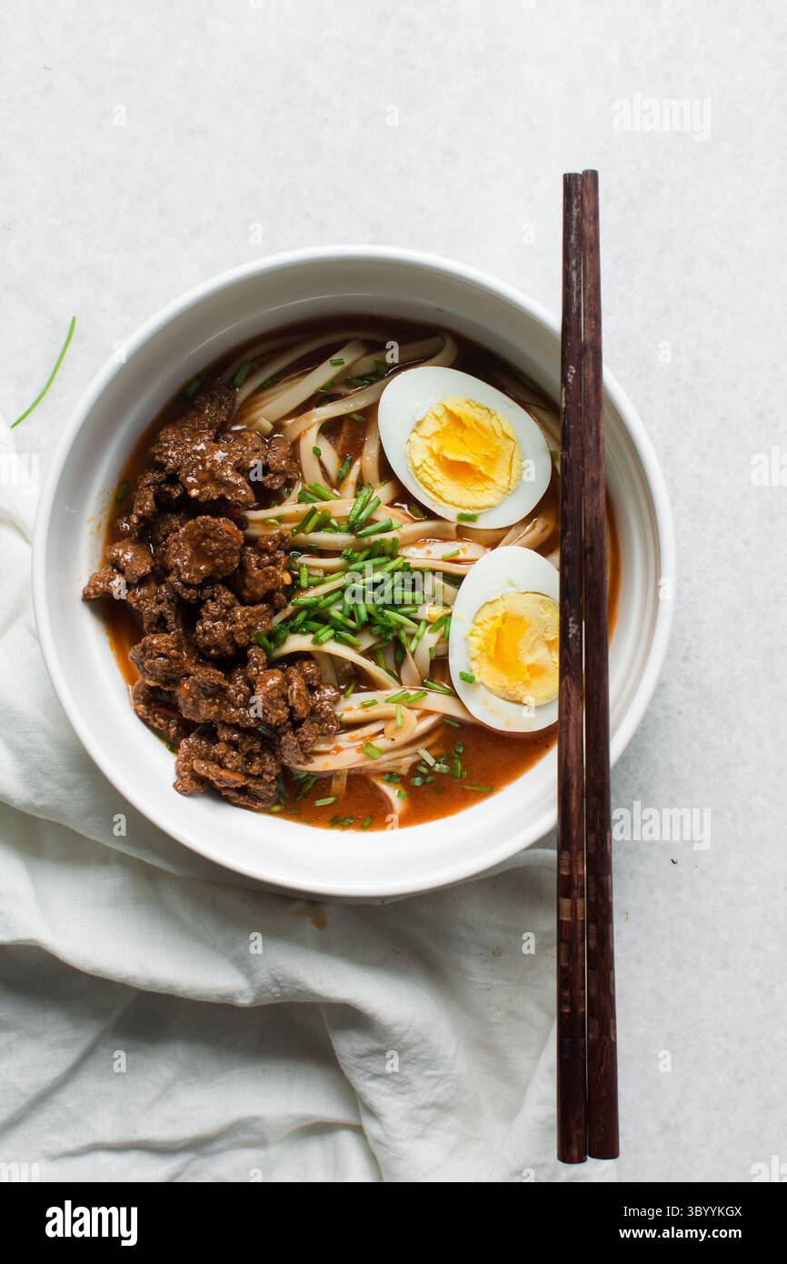 Blick von oben auf scharfe Nudelsuppe mit Eiern und Rindfleischstücken in einer weißen Schüssel, Blick von oben auf Nudeln Eier in einer scharfen Brühe mit Zwiebeln Stockfoto