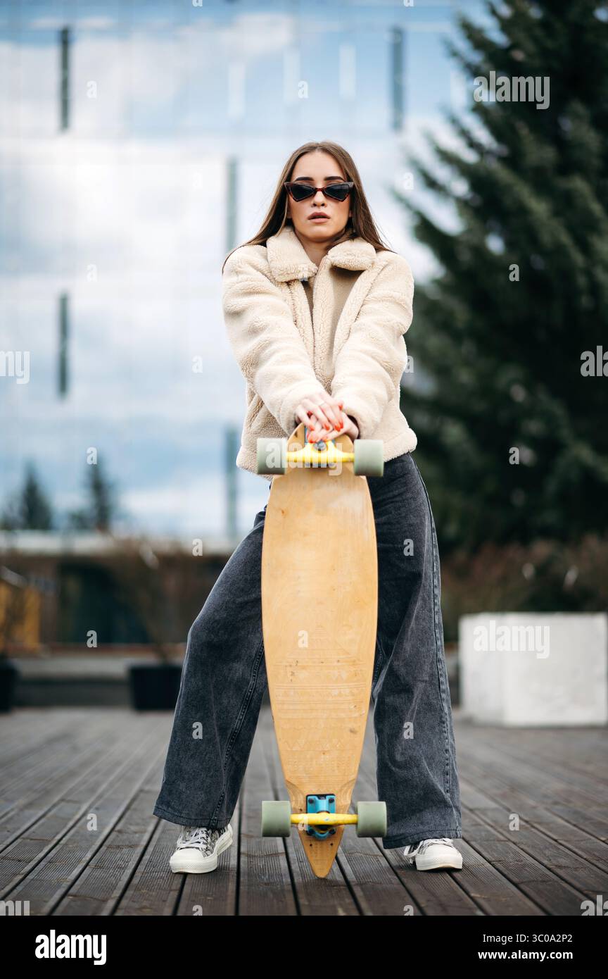 Junge Frau in stilvoller Kleidung posiert auf dem Dach mit Skateboard Stockfoto