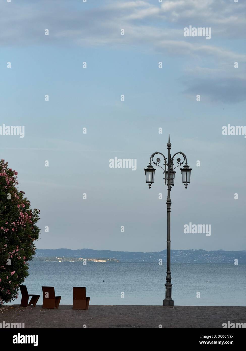 Lampenpfosten und Stühle an der Hafenfront von Lazise vor blauem Himmel. Gardasee, Italien Stockfoto