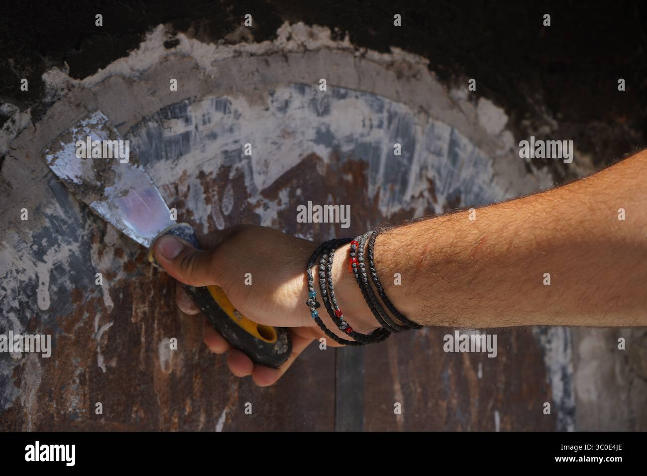 Die Hand eines jungen Mannes versiegelt eine holzbefeuerte Ofentür mit einem Spachtel und einer Aschemischung. Im Inneren warten Trays mit traditionellen kretischen Gerichten auf langsames Backen. Stockfoto