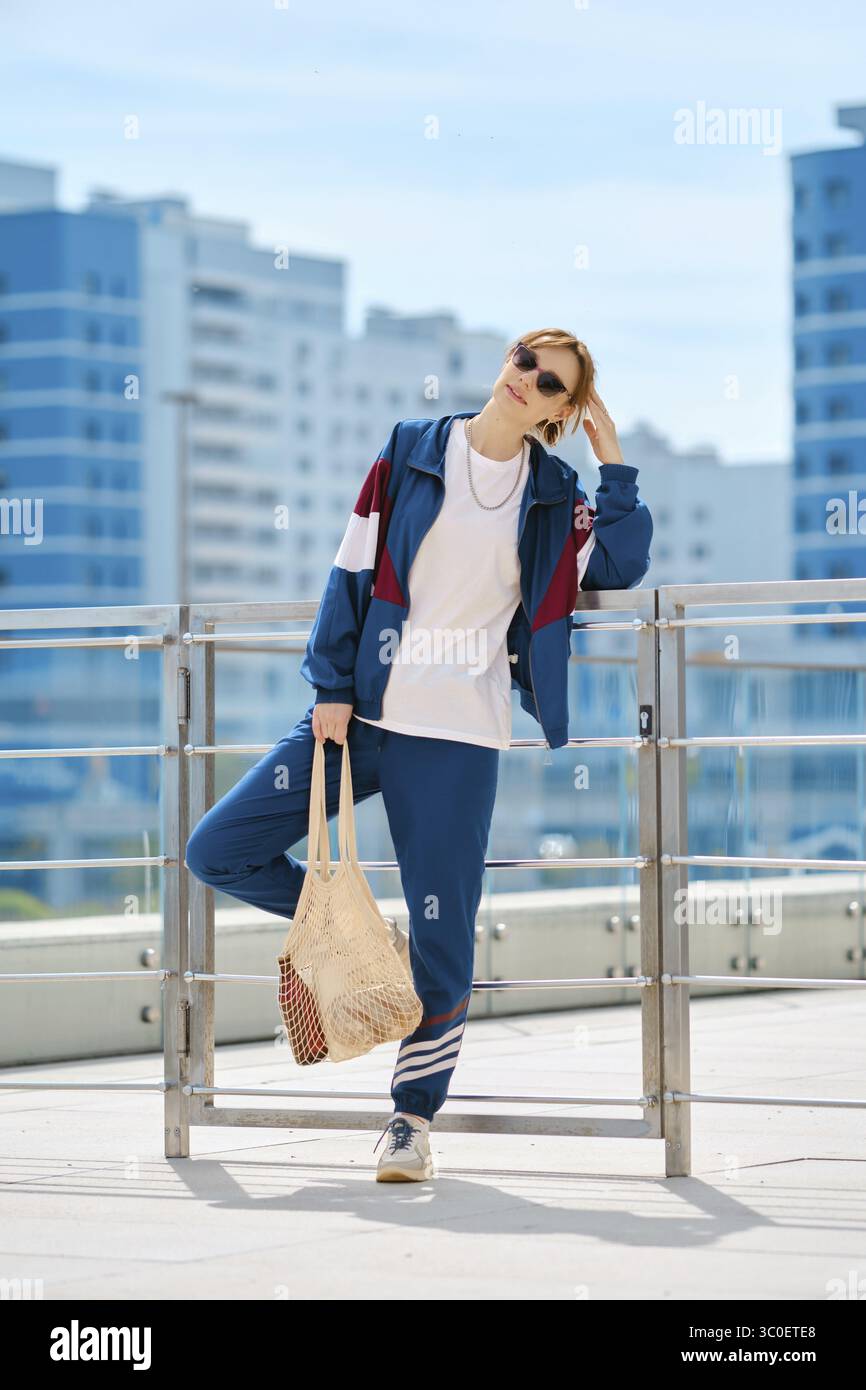 Eine Frau in sportlichem Outfit steht selbstbewusst auf einer Terrasse, umgeben von einem hellblauen Himmel und modernen Gebäuden Stockfoto