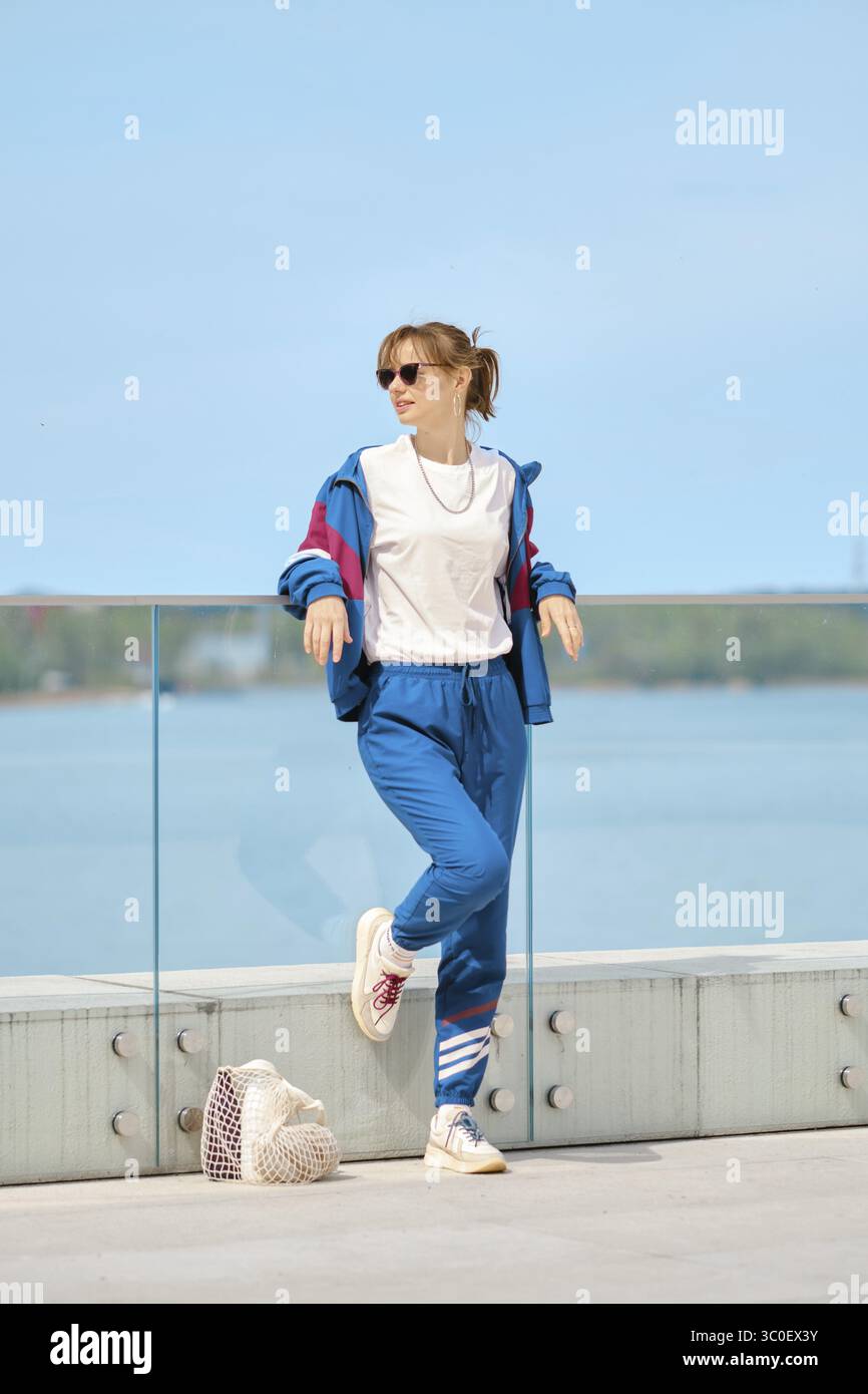 Eine Frau entspannt sich an einem Glasgeländer am Wasser an einem hellen sonnigen Tag. Sie trägt ein farbenfrohes sportliches Outfit und eine Sonnenbrille, die lässig und lässig wirkt Stockfoto