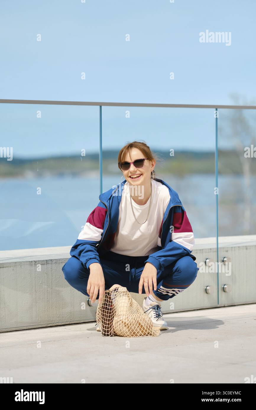 Ein junger Mensch hockt auf einer modernen Terrasse am malerischen Wasser, trägt eine Sonnenbrille und ein sportliches Outfit Stockfoto