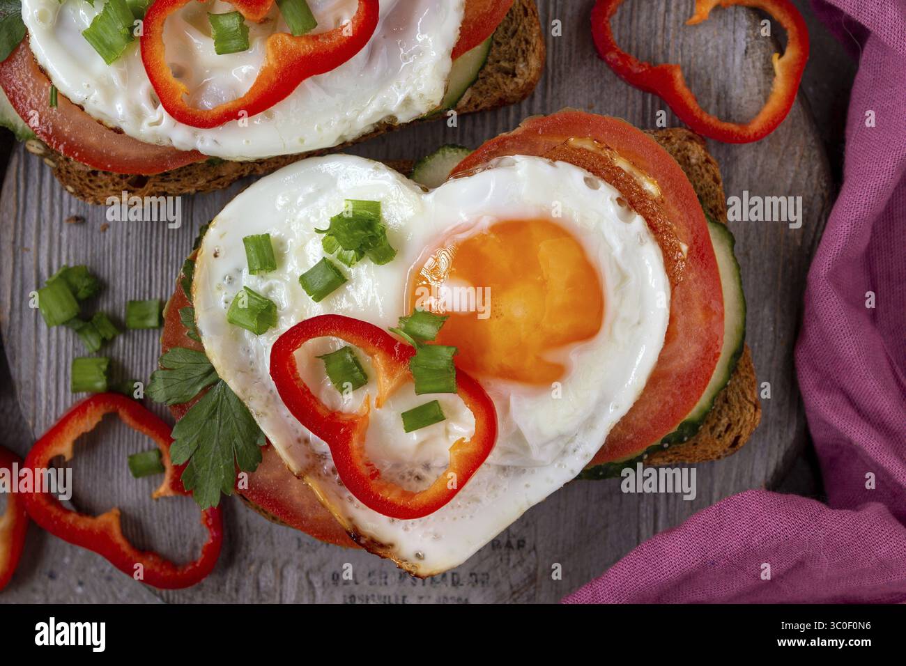 Sandwich mit Tomaten, Gurken und Ei in Herzform auf Holzbrett, lila Serviette Stockfoto