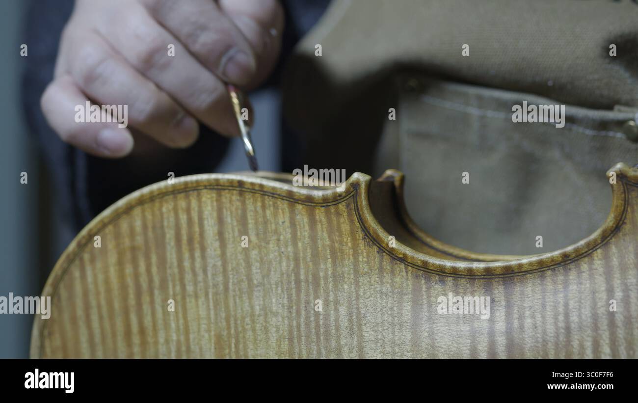 Eine Hand, die einen Pinsel in der Nähe einer Geige hält, mit sichtbarem Holzmaserung, Lackprozess der Geige, antikique Stockfoto