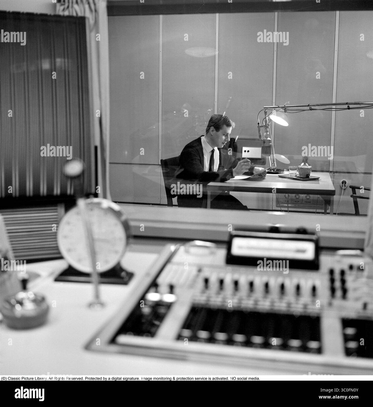 Radiostudio aus den 1960er Jahren. Radiomoderator Klas Burling (1941–2023) im Studio vor dem Mikrofon, wenn er auf Sendung ist, aus dem Raum der Radiomoduzenten und Tontechniker durch das schalldichte Fenster, das sie voneinander trennt, gesehen. 1963 Stockfoto