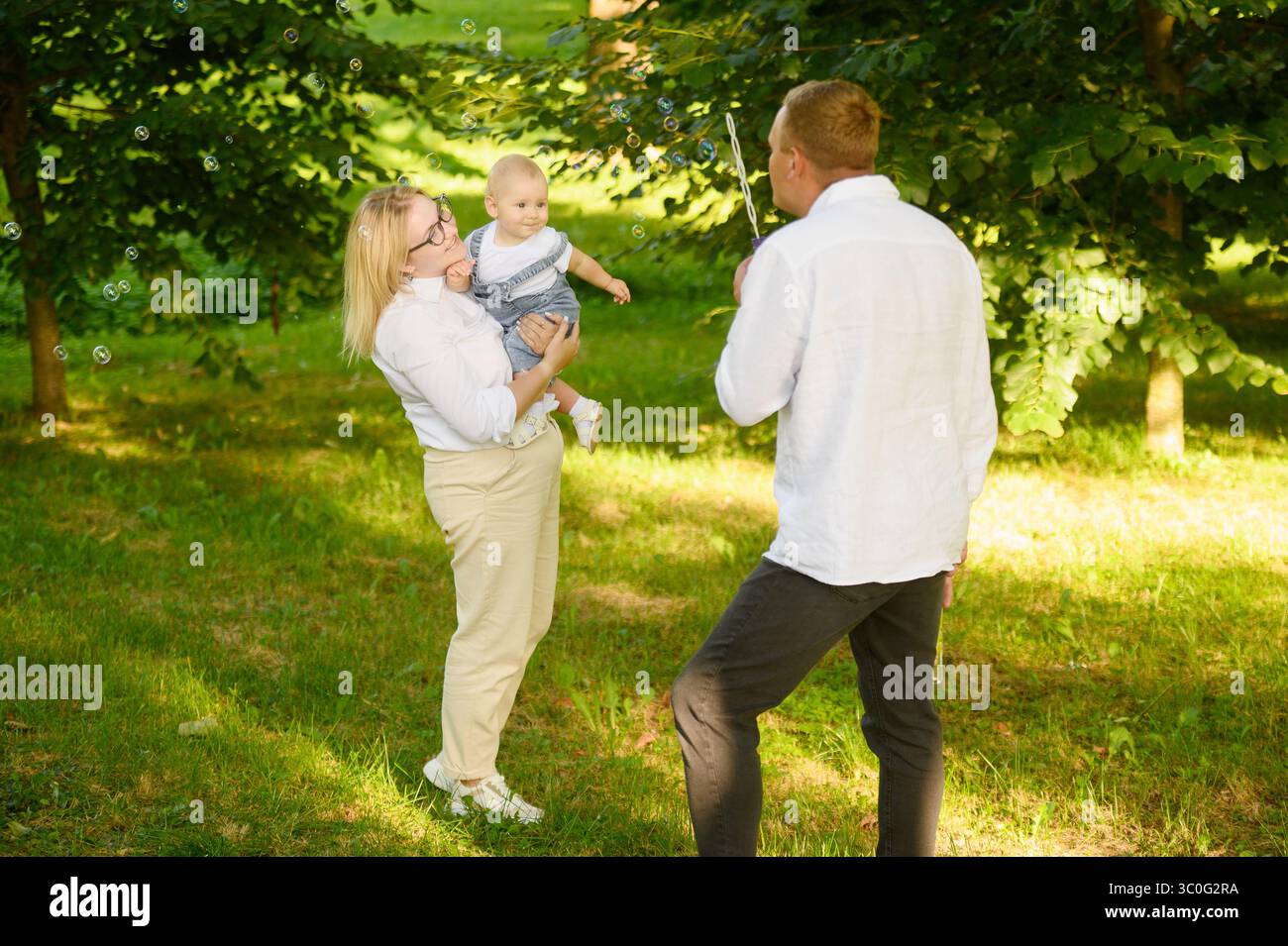 Glückliche Familie entspannt sich im Park. Vater spielt glücklich und bläst Seifenblasen, Mutter hält niedliches Baby in den Armen, lacht mit ihm, fängt Stockfoto